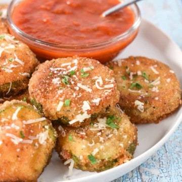 Breaded and fried zucchini rounds displayed on a white plate with a small bowl of red sauce in the background.
