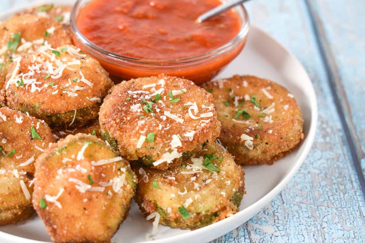 Breaded and fried zucchini rounds displayed on a white plate with a small bowl of red sauce in the background.
