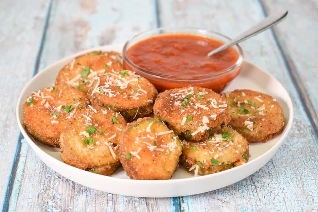 Fried zucchini rounds arranged on a white plate with a small bowl of marinara sauce.