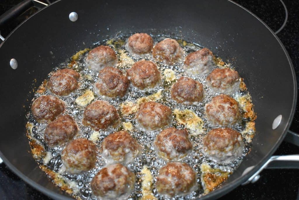 Browned small meatballs frying in oil in a large, black skillet.