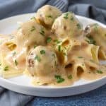 Small meatballs smothered in a light colored gravy served on a bed of egg noodles on a white plate.