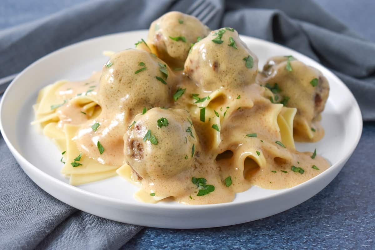 Small meatballs smothered in a light colored gravy served on a bed of egg noodles on a white plate.