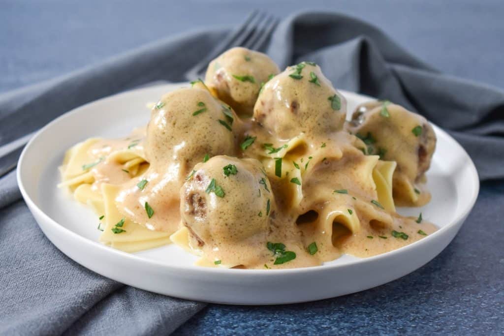 Small meatballs smothered in a light colored gravy served on a bed of egg noodles on a white plate.