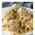 Small meatballs smothered in a light colored gravy served on a bed of egg noodles on a white plate.