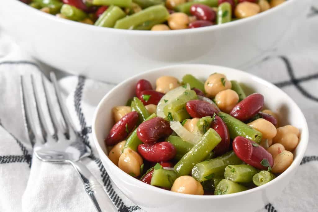 Three bean salad served in a small white bowl with a larger bowl in the background displayed on a white cloth with black checkered lines.