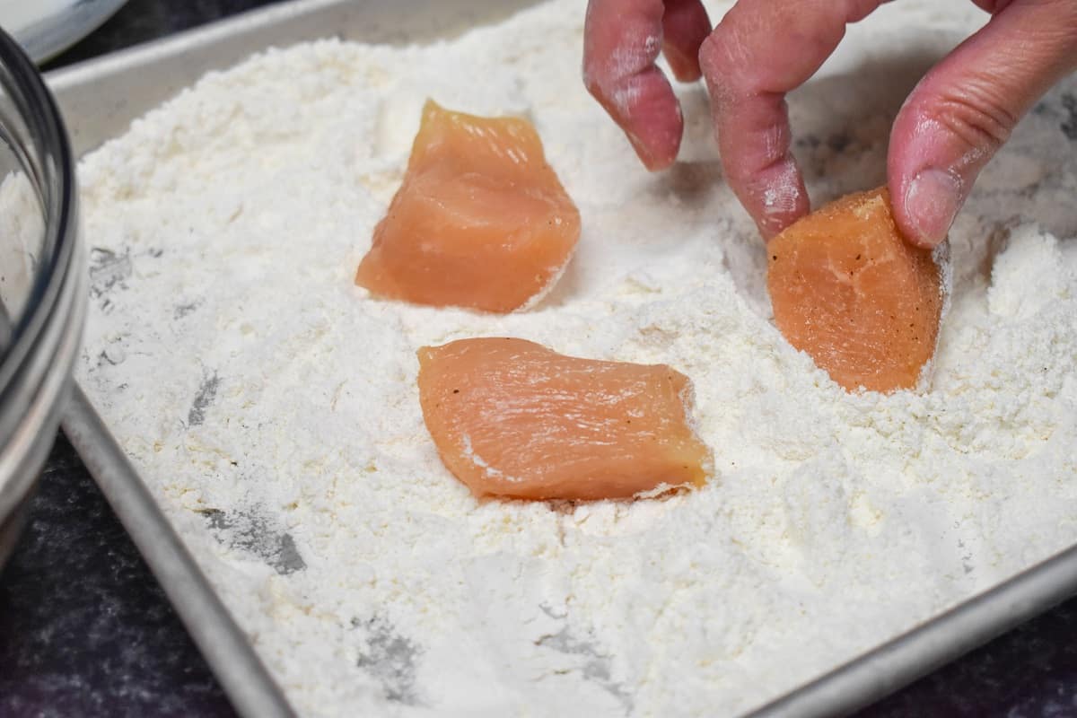 Three small pieces of raw chicken being coated in flour.