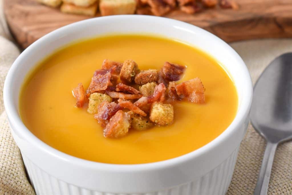 Creamy butternut squash soup served in a large, white ramekin and garnished with bacon bits and croutons.