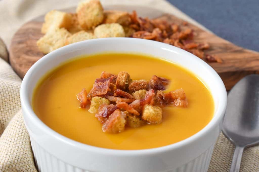 Creamy butternut squash soup served in a large, white ramekin and garnished with bacon bits and croutons.