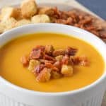 Creamy butternut squash soup served in a large, white ramekin and garnished with bacon bits and croutons.