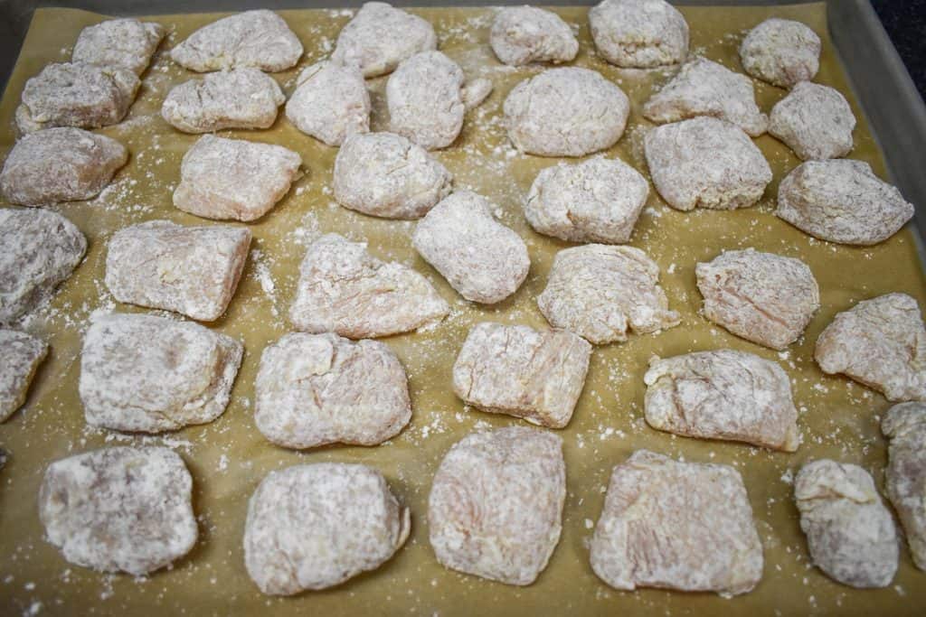 Small pieces of chicken breasts coated in flour and displayed on a large, parchment lined baking sheet.