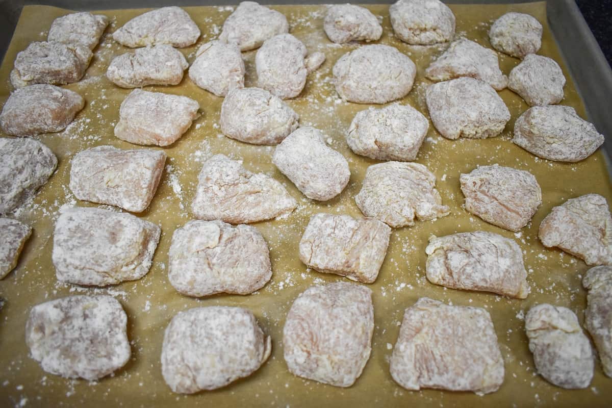 Small pieces of chicken breasts coated in flour and displayed on a large, parchment lined baking sheet.