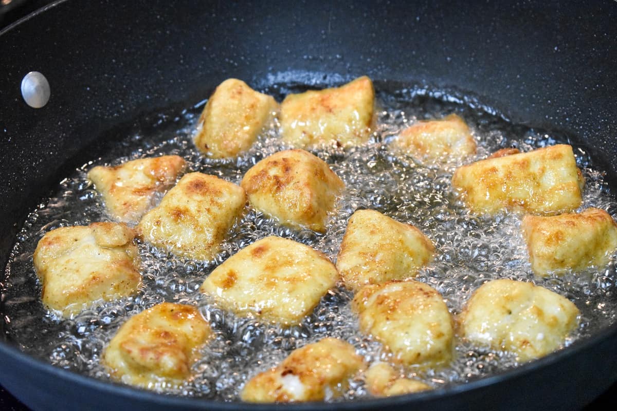 Flour coated chicken pieces frying in oil in a large, black skillet.