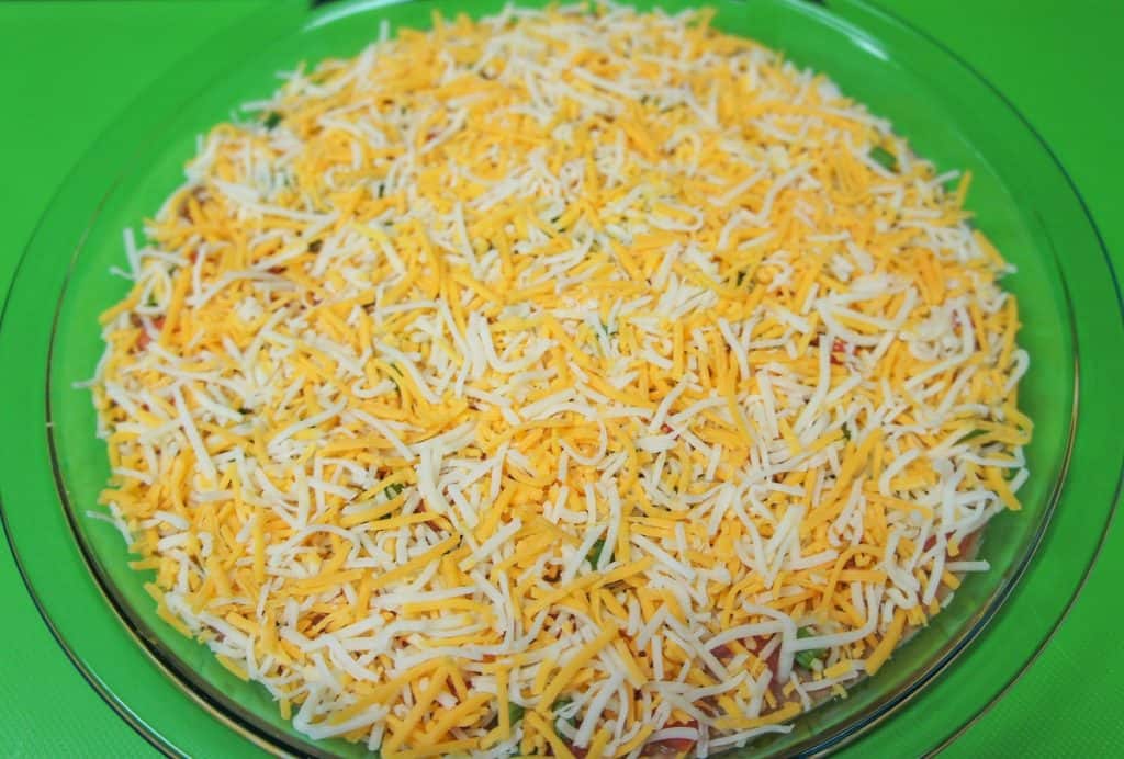 A layer of finely shredded cheese covering refried beans and diced tomatoes and green onions in a glass pie plate.