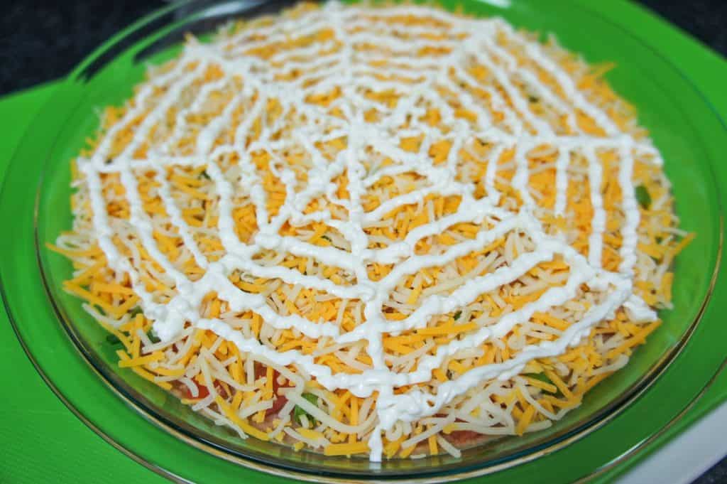 Sour cream lines made into a spider web on the cheese covered bean dip in a glass pie plate.