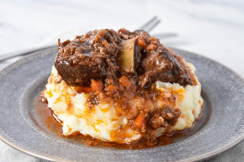 Short ribs and sauce served on a bed of mashed potatoes on a gray plate.
