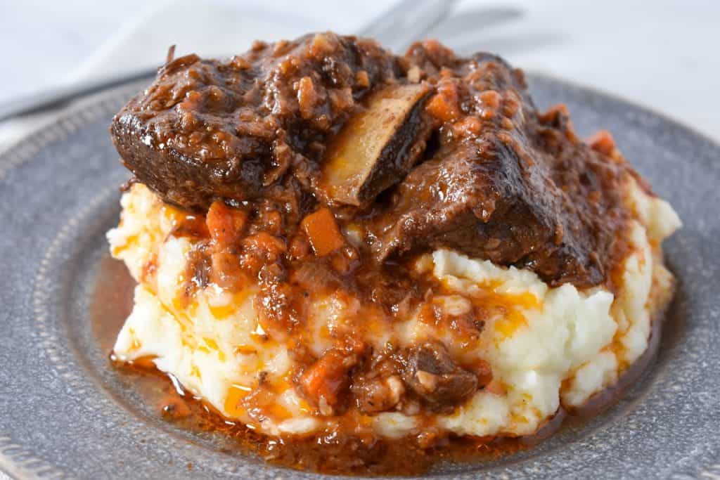 A close up picture of short ribs and sauce served on a bed of mashed potatoes on a gray plate.