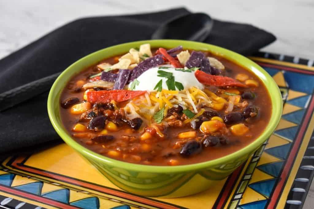 Turkey taco soup served in a green bowl and set on a colorful southwestern style plated with a black napkin and spoon in the background.