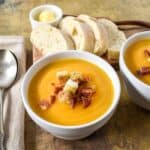 Bowl of creamy butternut squash soup topped with crispy bacon and croutons, served with sliced bread and butter on the side.