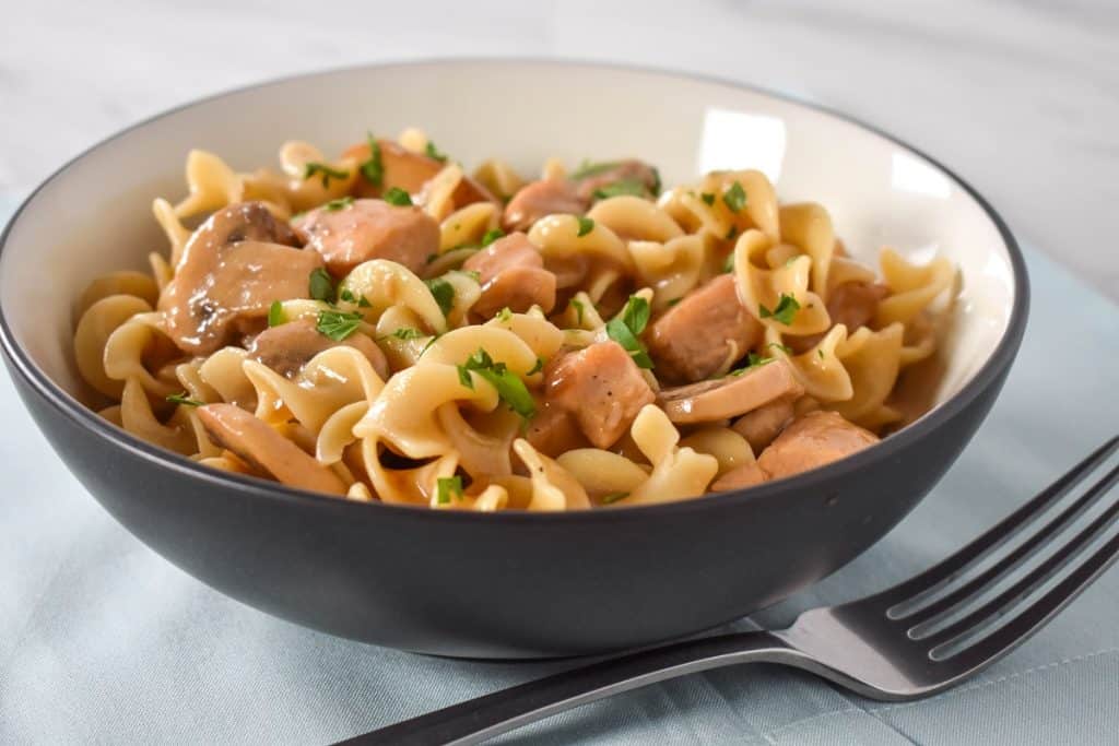 Egg noodles, cubed chicken and sliced mushrooms served in a white bowl on a light blue linen.