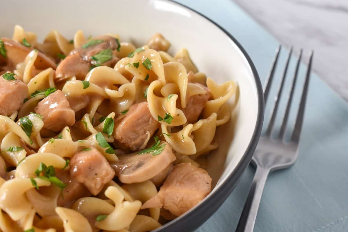 Egg noodles, cubed chicken and sliced mushrooms served in a white bowl on a light blue linen.