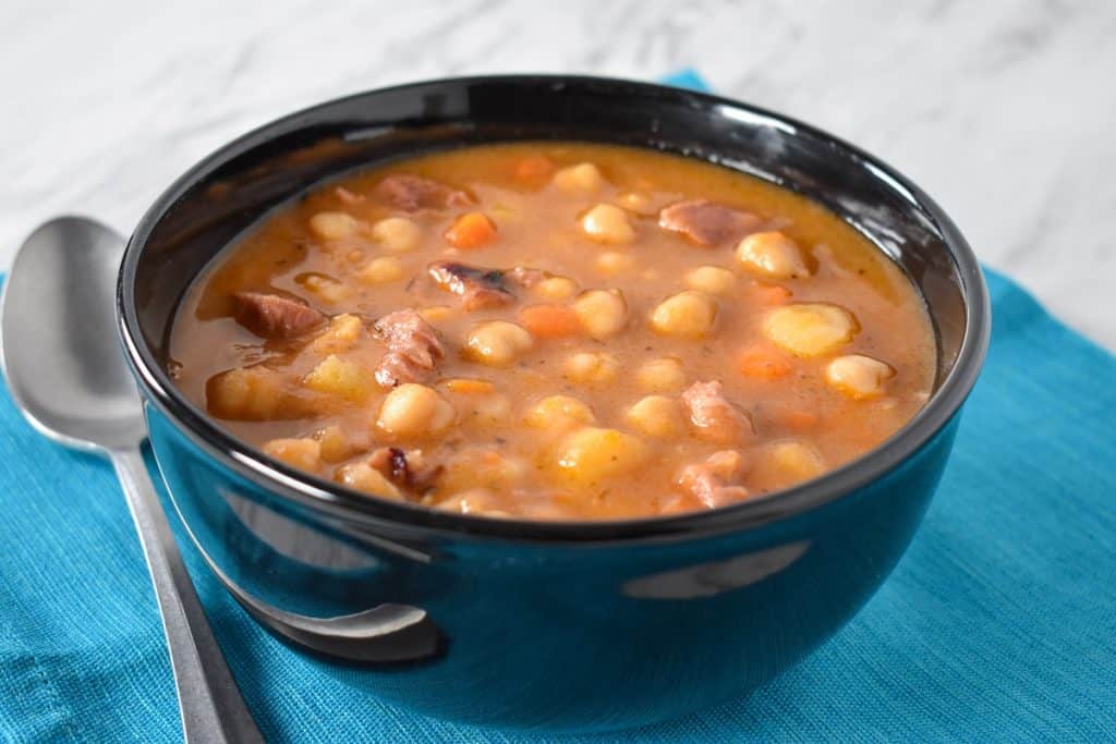 Chickpeas soup served in a black bowl, set on an aqua colored linen with a spoon on the side.