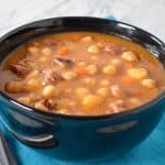 Chickpeas soup served in a black bowl, set on an aqua colored linen with a spoon on the side.
