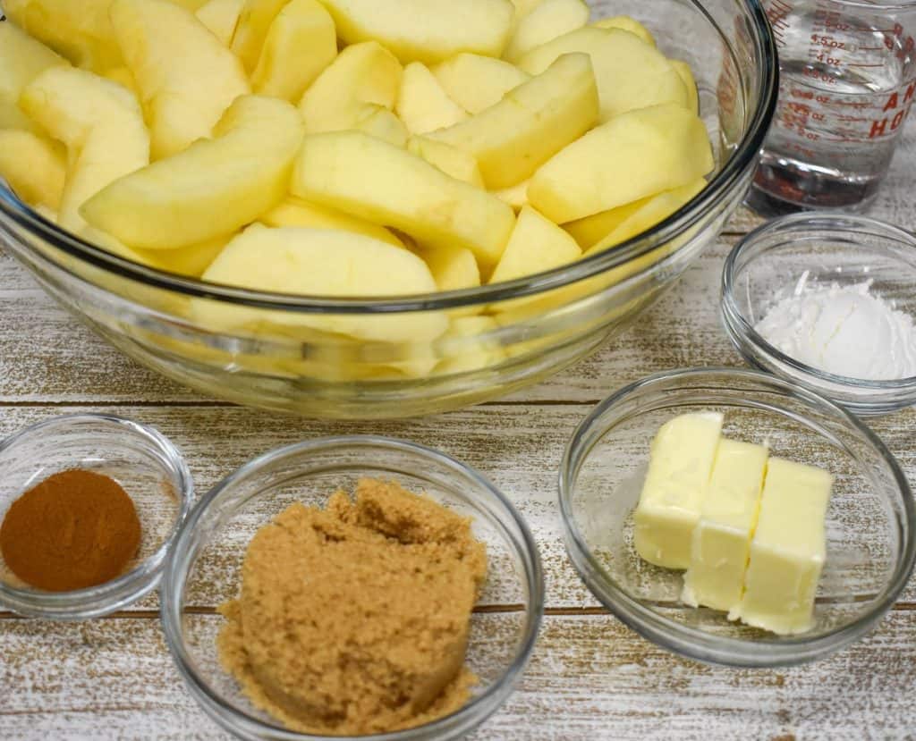 Sliced, peeled apples in a large bowl with cinnamon, brown sugar, butter and corn starch in smaller bowls.