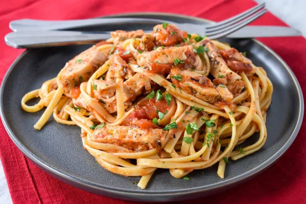 Linguine noodles tossed with tomatoes and sliced chicken breast served on a gray plate on a red cloth napkin.