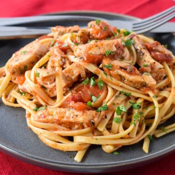 Linguine noodles tossed with tomatoes and sliced chicken breast served on a gray plate on a red cloth napkin.