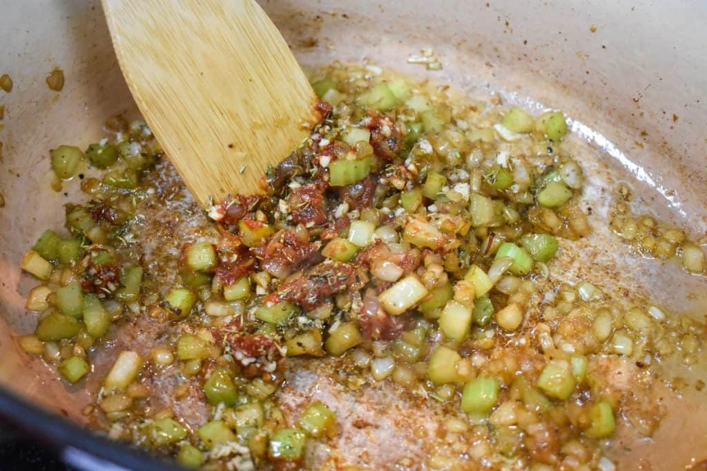 Diced onion, celery, minced garlic, tomato paste and spices being cooked in a large pot.