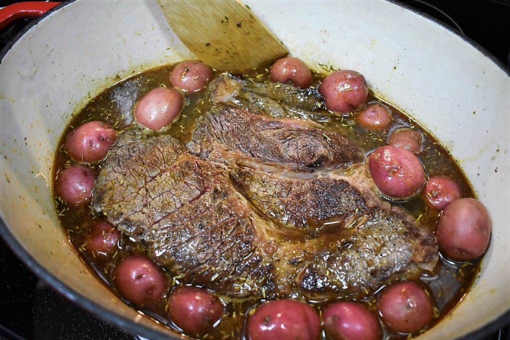A large browned pot roast in a large pot with potatoes arranged around it.