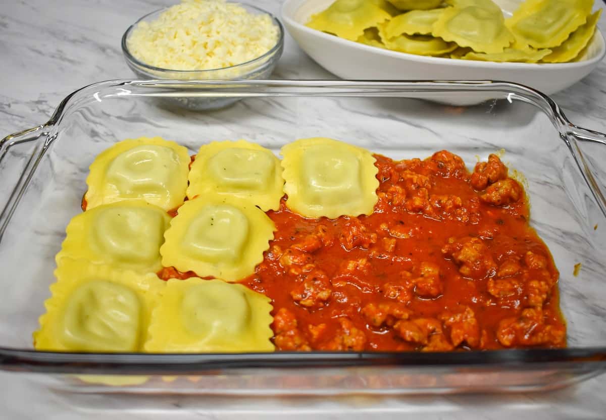 Ravioli being layered on sausage pasta sauce in a glass casserole dish.