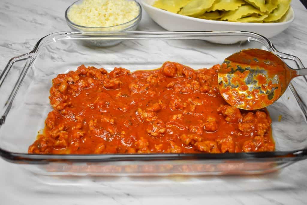 A layer of Italian sausage pasta sauce being added to a glass casserole dish.