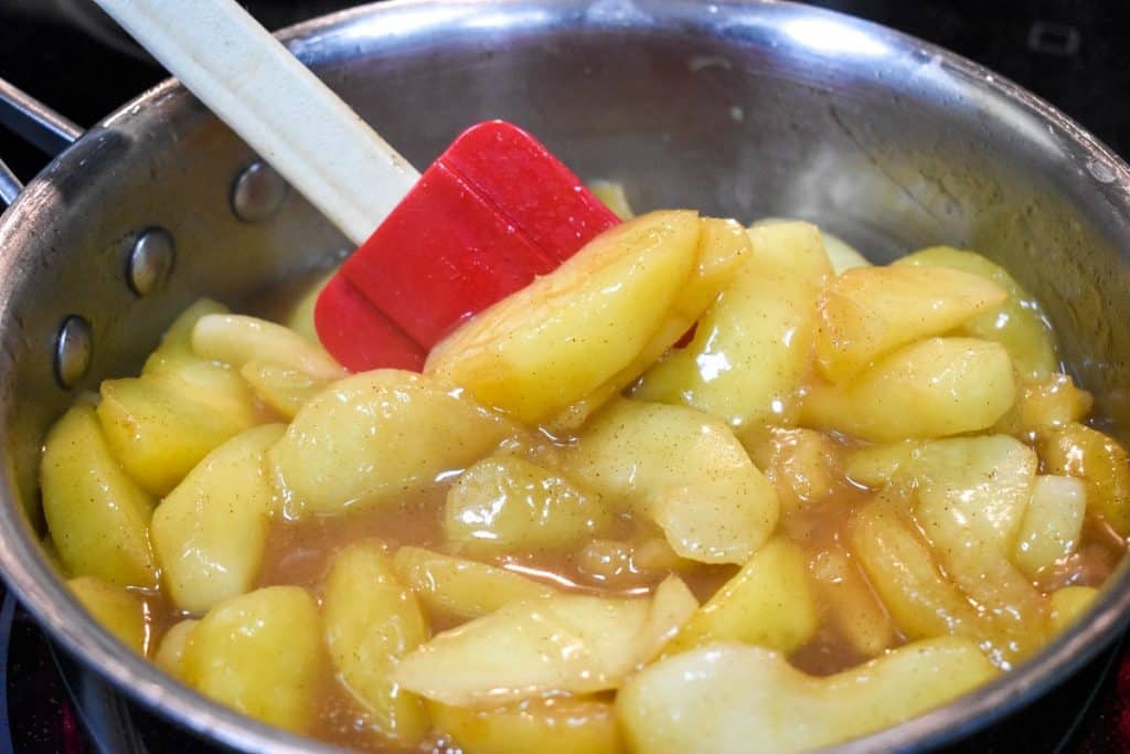 Cinnamon apples in a saucepan being stirred by a red spatula.