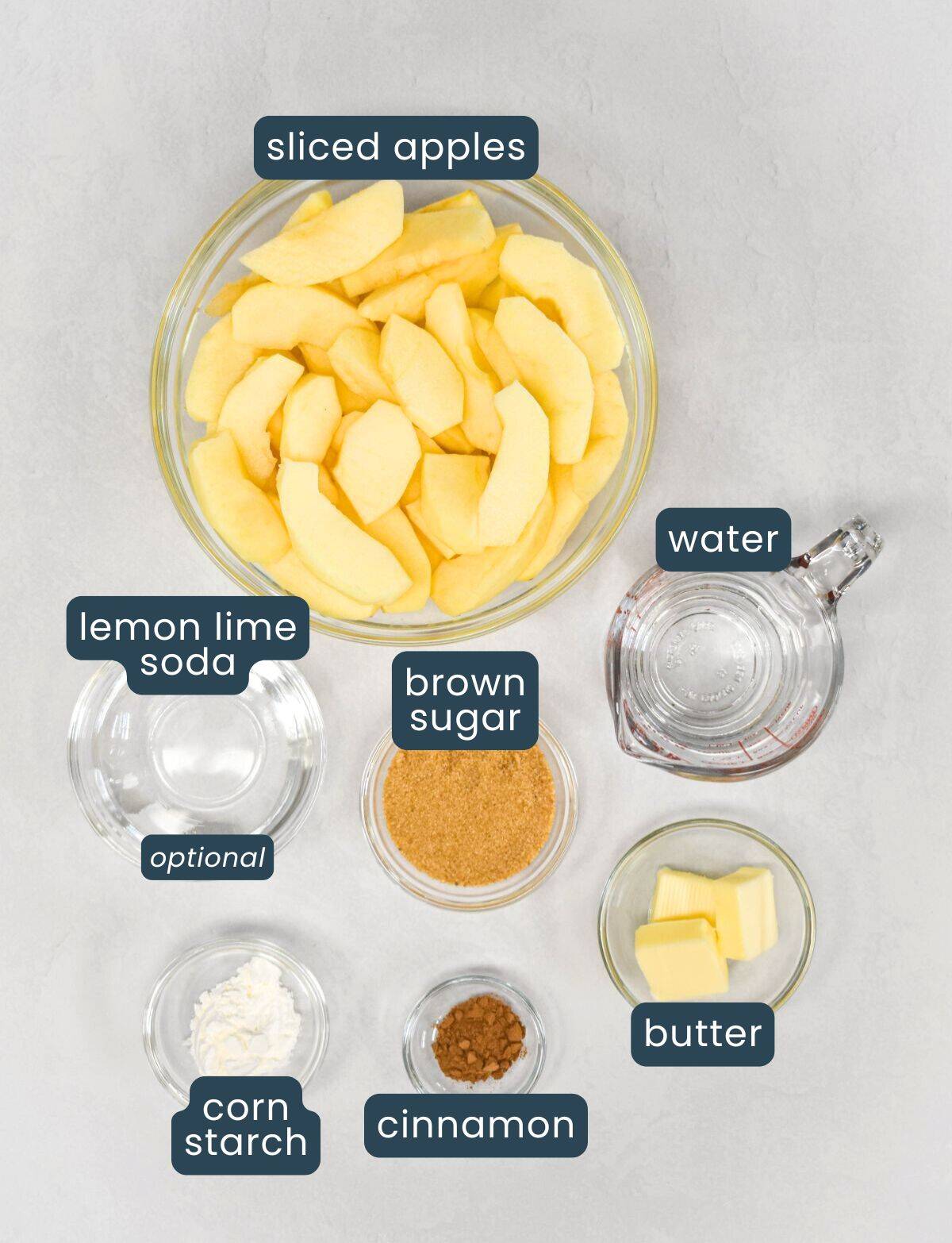 Ingredients for cinnamon apples arranged on a light surface, including sliced apples, brown sugar, butter, cinnamon, cornstarch, water, and lemon-lime soda.