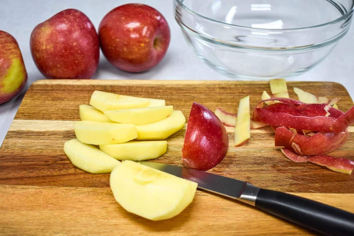 Apples being peeled and sliced on a wooden cutting board with a paring knife and an empty glass bowl in the background.