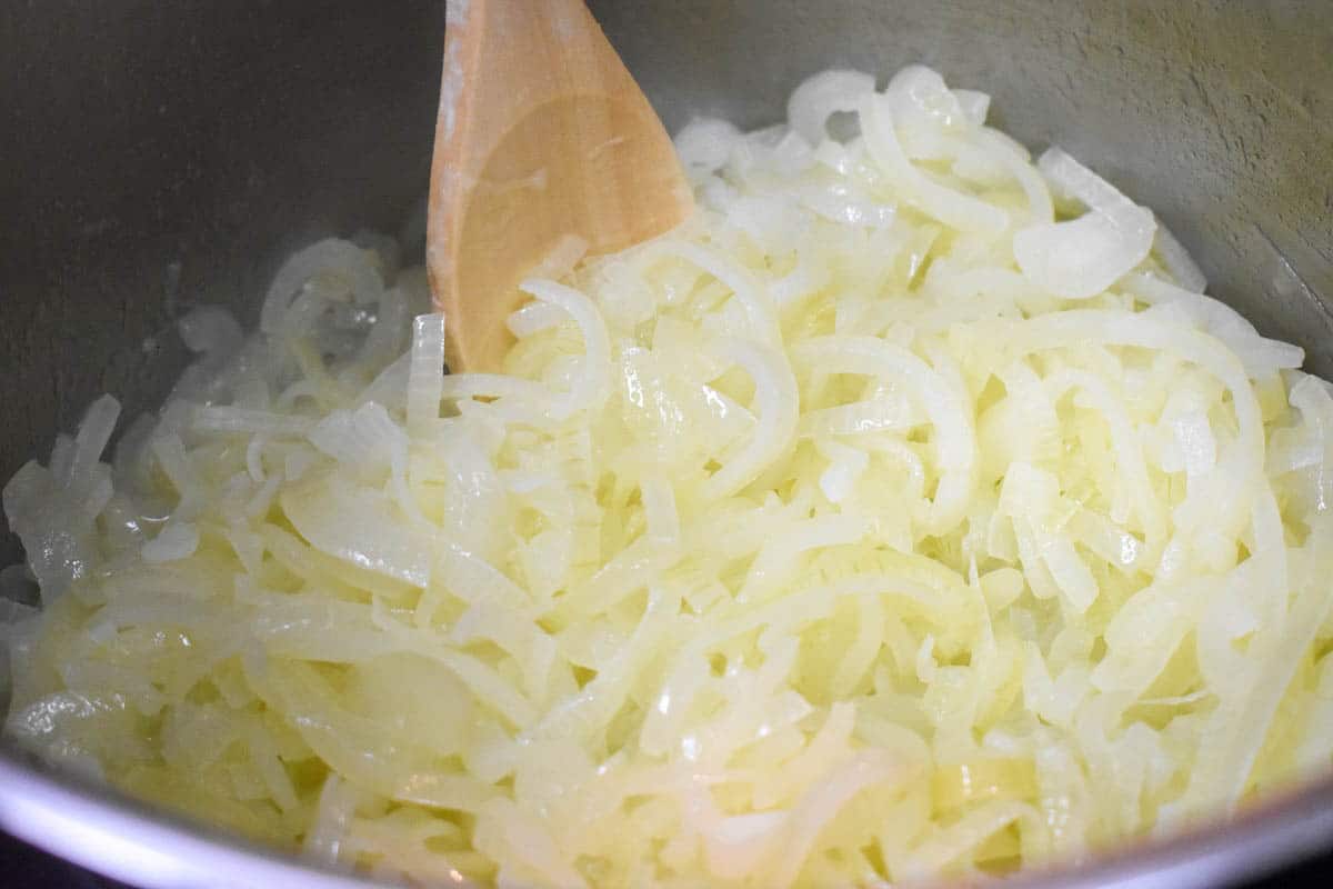 An image of sliced onions starting to soften in a large pot with a wooden spoon.