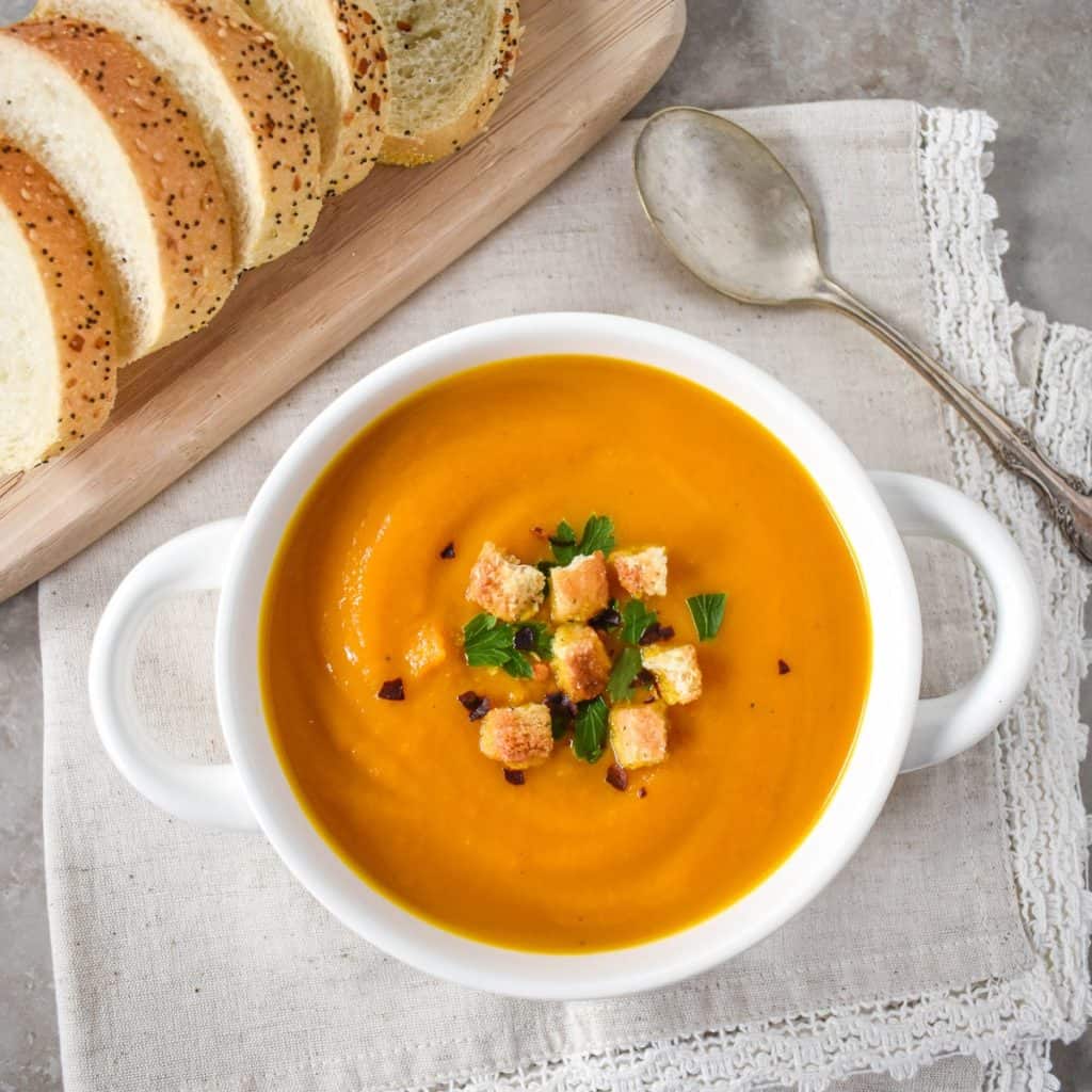 The carrot ginger soup served in a white bowl and garnished with chopped parsley, croutons, and red pepper flakes. The bowl is set on a light beige linen with a spoon to the right side and sliced bread on a wood cutting board to the top left.