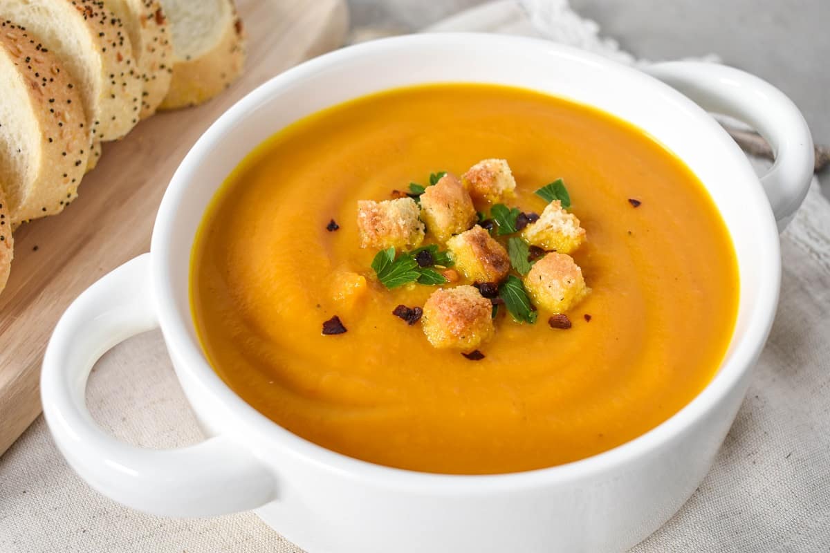 A close up of the finished carrot ginger soup served in a white bowl and garnished with croutons, chopped parsley, and red pepper flakes. 