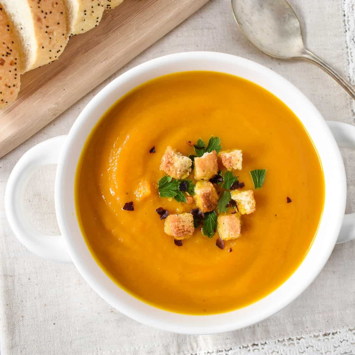 The carrot ginger soup served in a white bowl and garnished with chopped parsley, croutons, and red pepper flakes. The bowl is set on a light beige linen with a spoon to the right side and sliced bread on a wood cutting board to the top left.