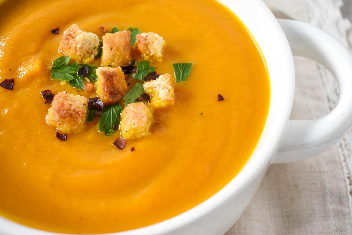 A close up of the finished carrot ginger soup served in a white bowl and garnished with croutons, chopped parsley, and red pepper flakes.