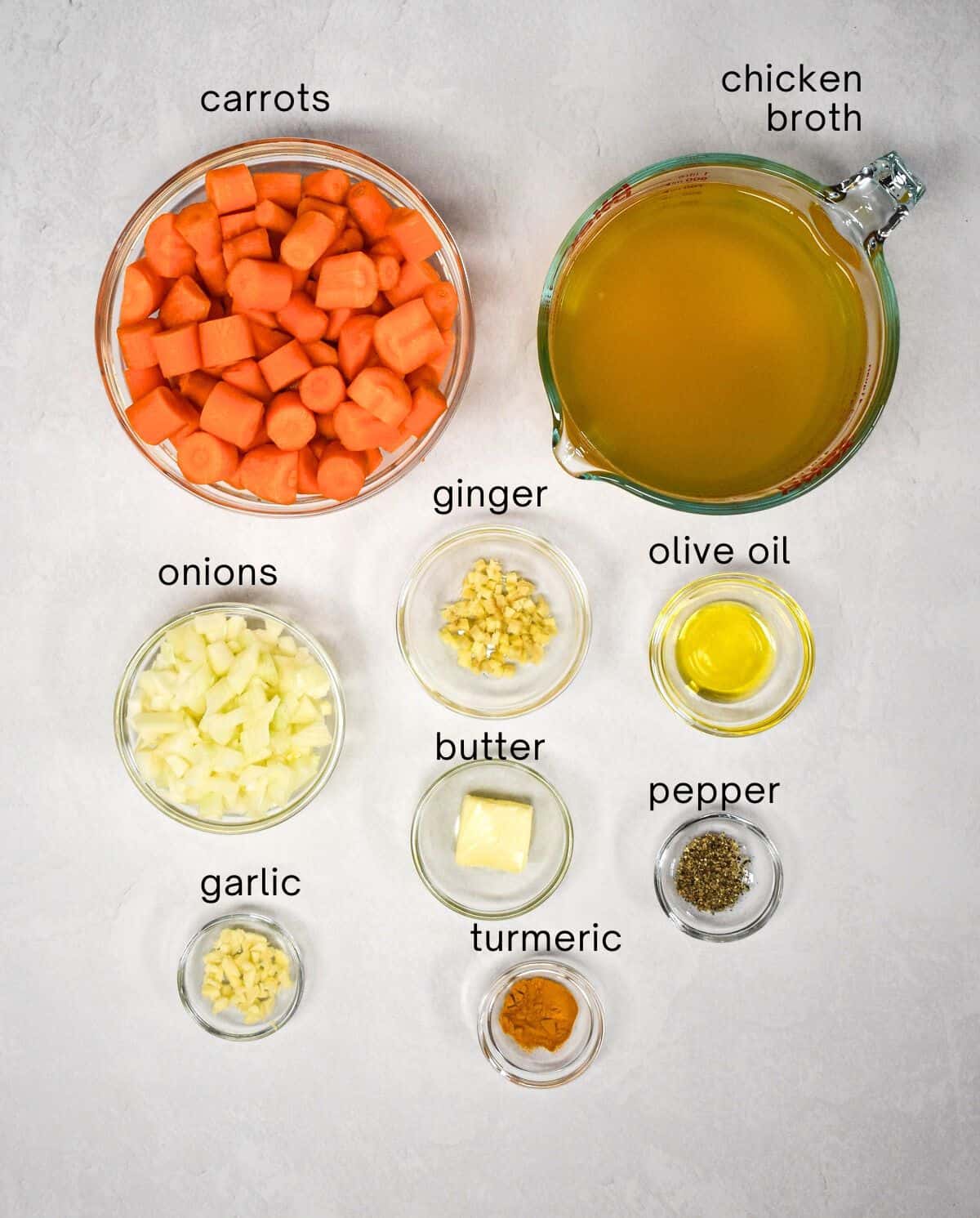 The ingredients for the soup prepped and arranged in glass bowls on a white table. Each ingredient is labeled with small. black letters.