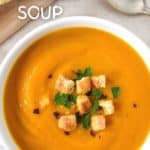 The carrot ginger soup served in a white bowl and garnished with chopped parsley, croutons, and red pepper flakes. The bowl is set on a light beige linen with a spoon to the right side and sliced bread on a wood cutting board to the top left. There is an overlay on the picture with the title in white letters and creamy soup on the bottom.