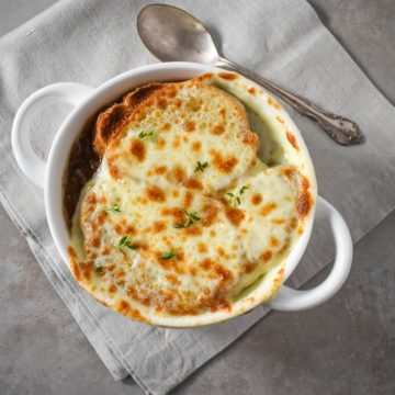 An image of the French onion soup served in a white soup bowl and set on a beige linen.