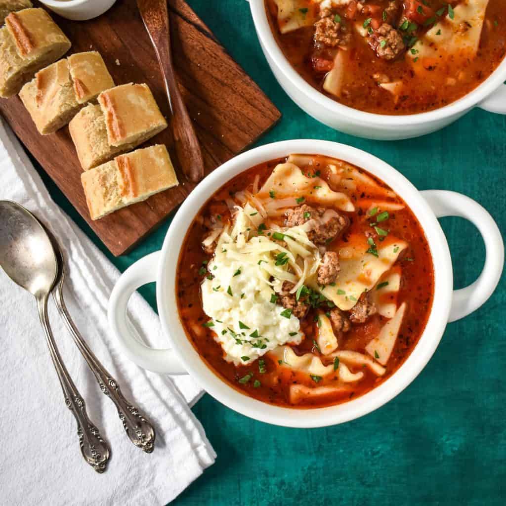 The lasagna soup served in white bowls with sliced bread and spoons to the left side.