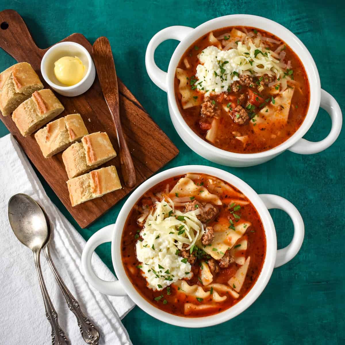 Two bowls of the finished soup set on a green table with sliced bread, butter and spoons to the left hand side.