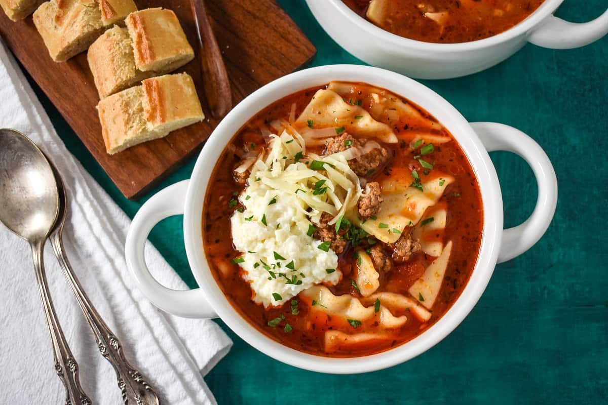 The lasagna soup served in white bowls with sliced bread and spoons to the left side.