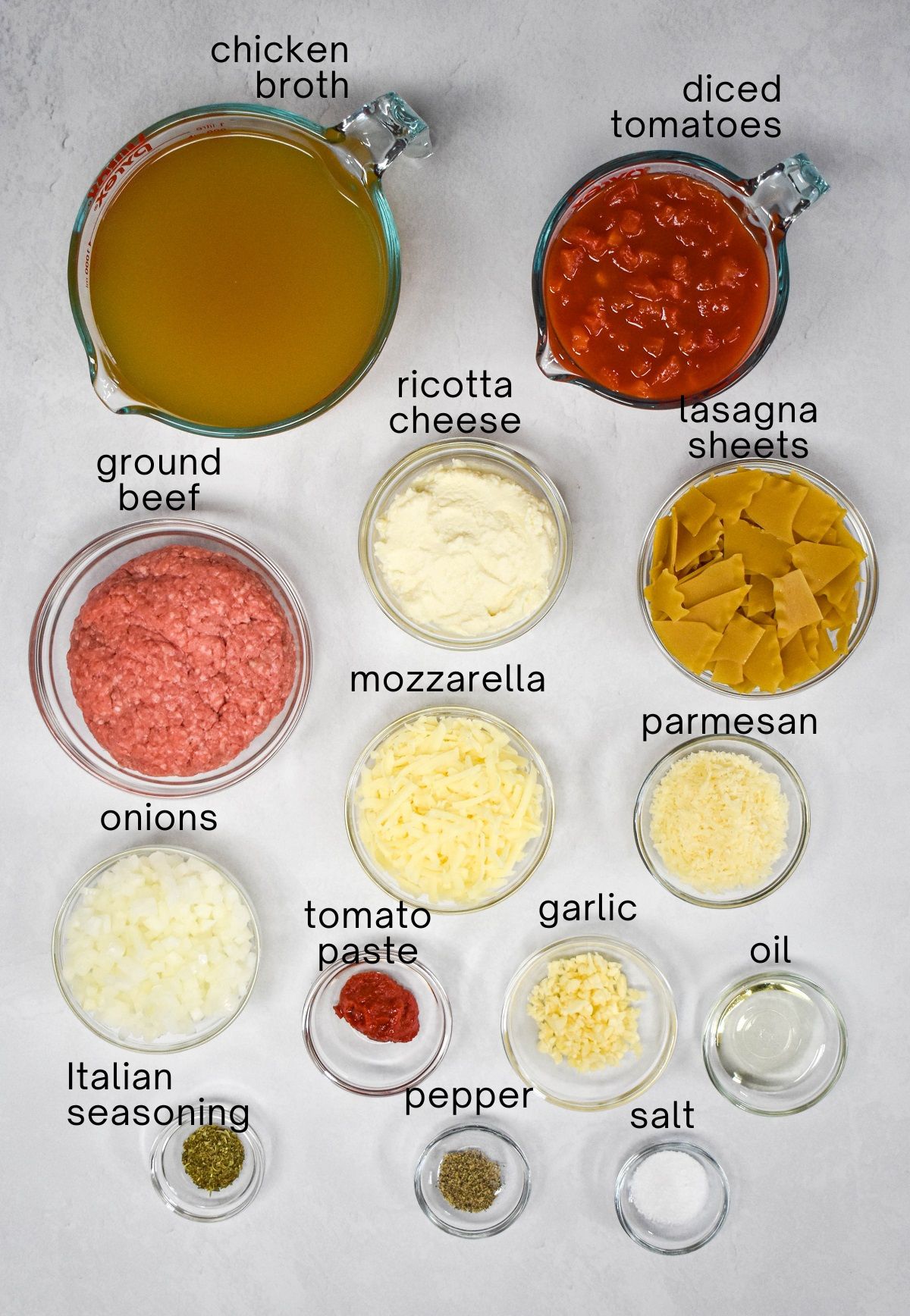 The ingredients for the lasagna soup prepped and arranged in glass bowls set on a white table, with each labeled with small, black letters.