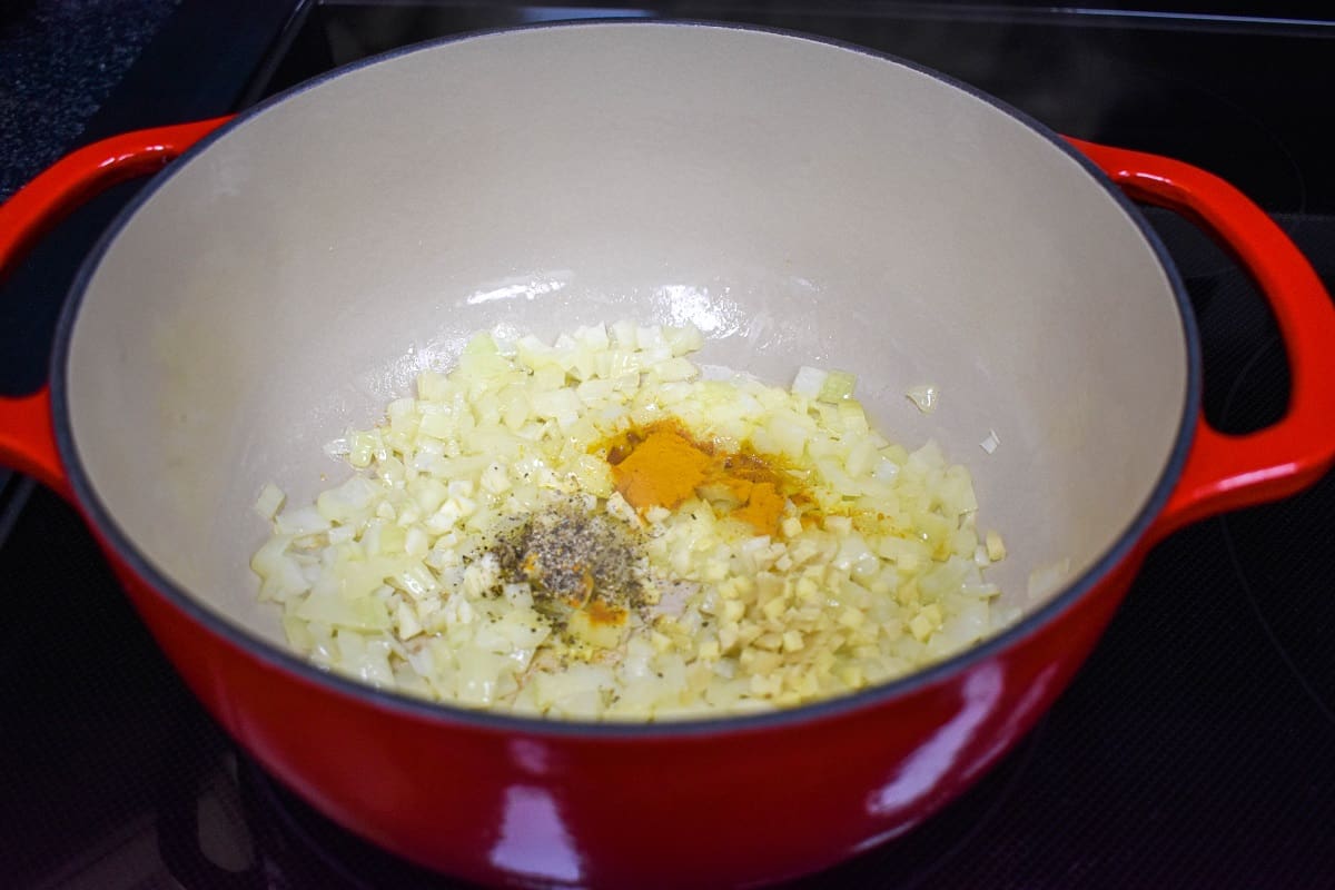 Minced garlic, ginger, turmeric, and black pepper added to the diced onions cooking in a large red and white pot.