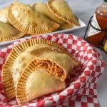 Chicken empanadas in a basket lined with a red and white checkered paper with hot sauce and a platter of empanadas in the background.